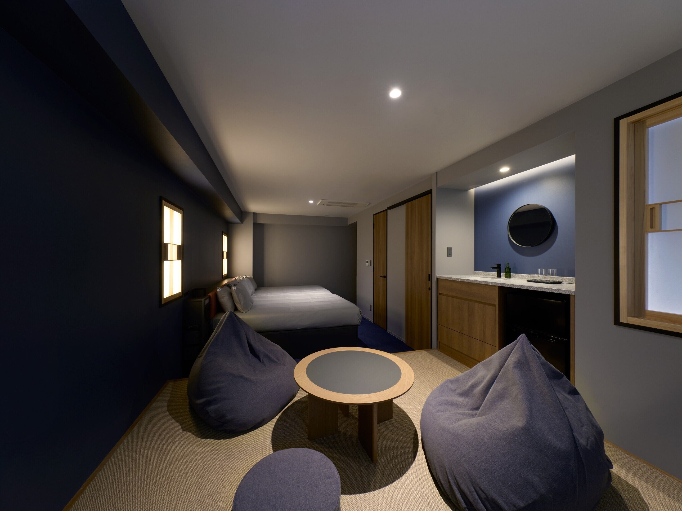 A modern hotel room with a bed, a round table, bean bags, a small kitchen area, and a bathroom with a mirror and sink.