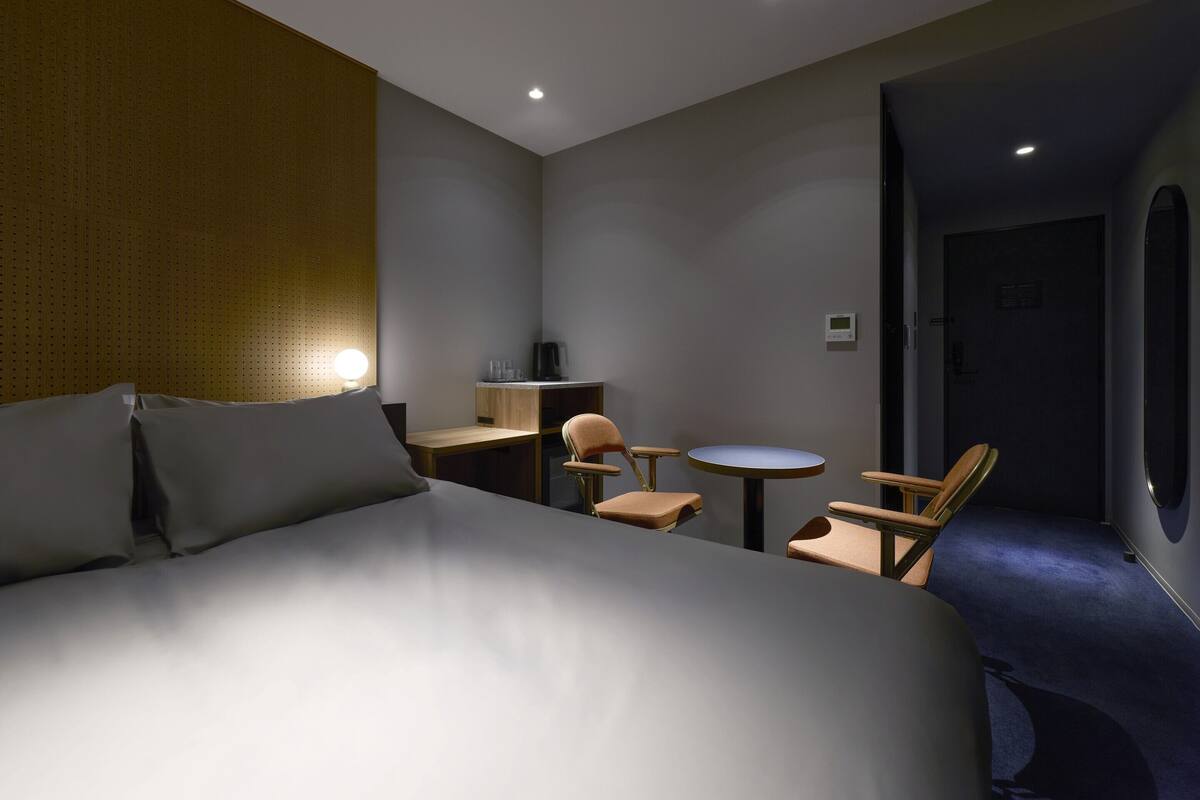 A hotel room with a bed, a desk, a chair, a small table, and a door.