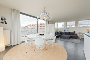 Ein modernes Wohnzimmer mit einem runden Esstisch, weißen Stühlen, einem grauen Ecksofa und einem großen Fenster mit Blick auf die Stadt.