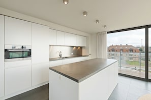 Eine moderne Küche mit weißen Schränken, einer großen Kochinsel und Blick auf Gebäude durch große Fenster.
