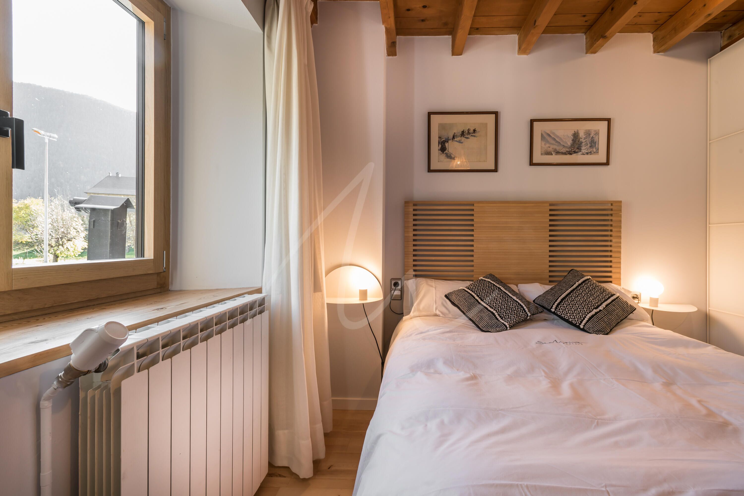 Een slaapkamer met een houten hoofdbord, twee wandlampen, een radiator en een raam met gordijnen.