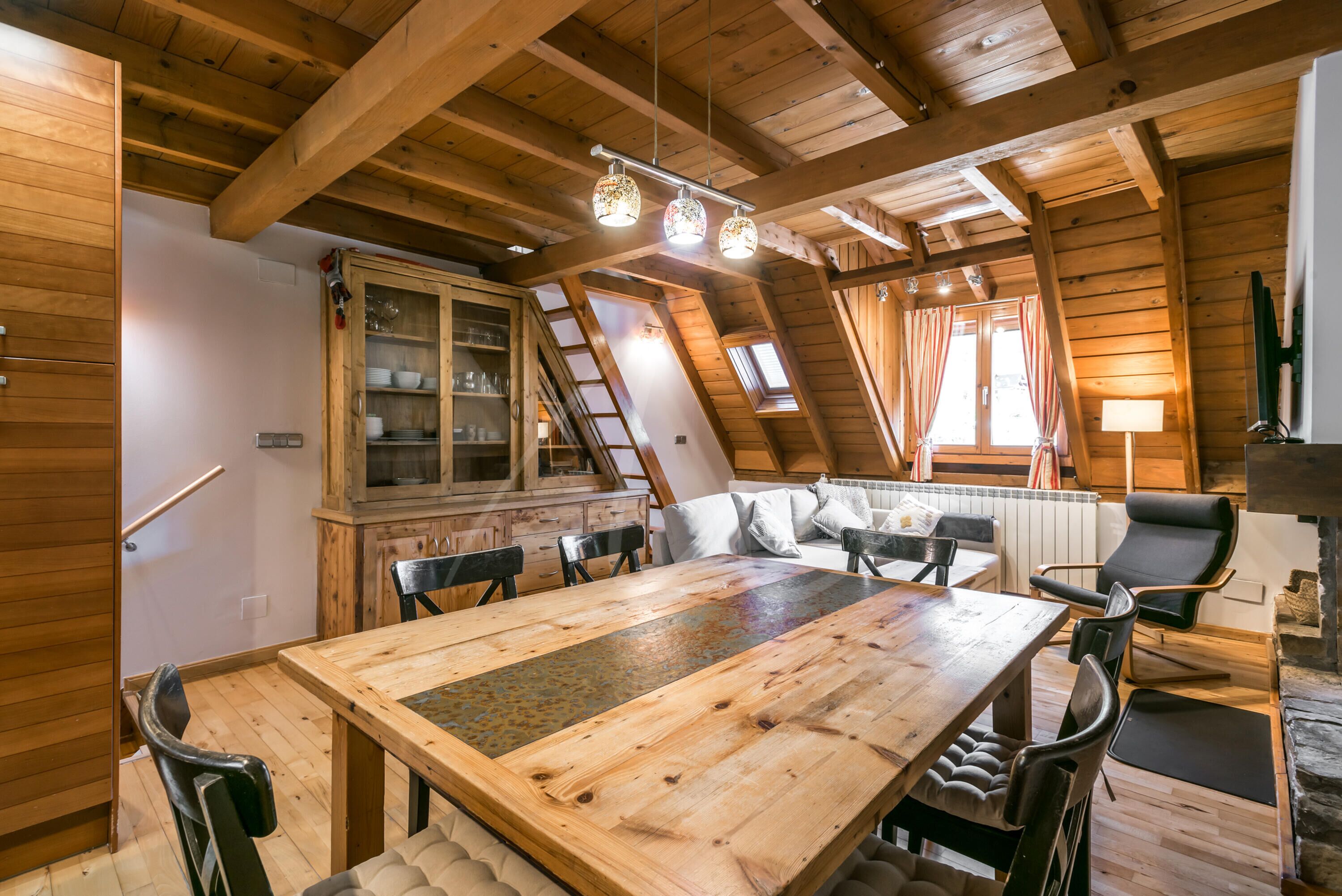 Een houten eethoek met een grote tafel en stoelen, een kast met glazen deuren, en een gezellige zithoek met kussens.