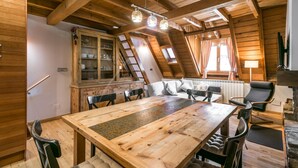 Een houten eethoek met een grote tafel en stoelen, een kast met glazen deuren, en een gezellige zithoek met kussens.