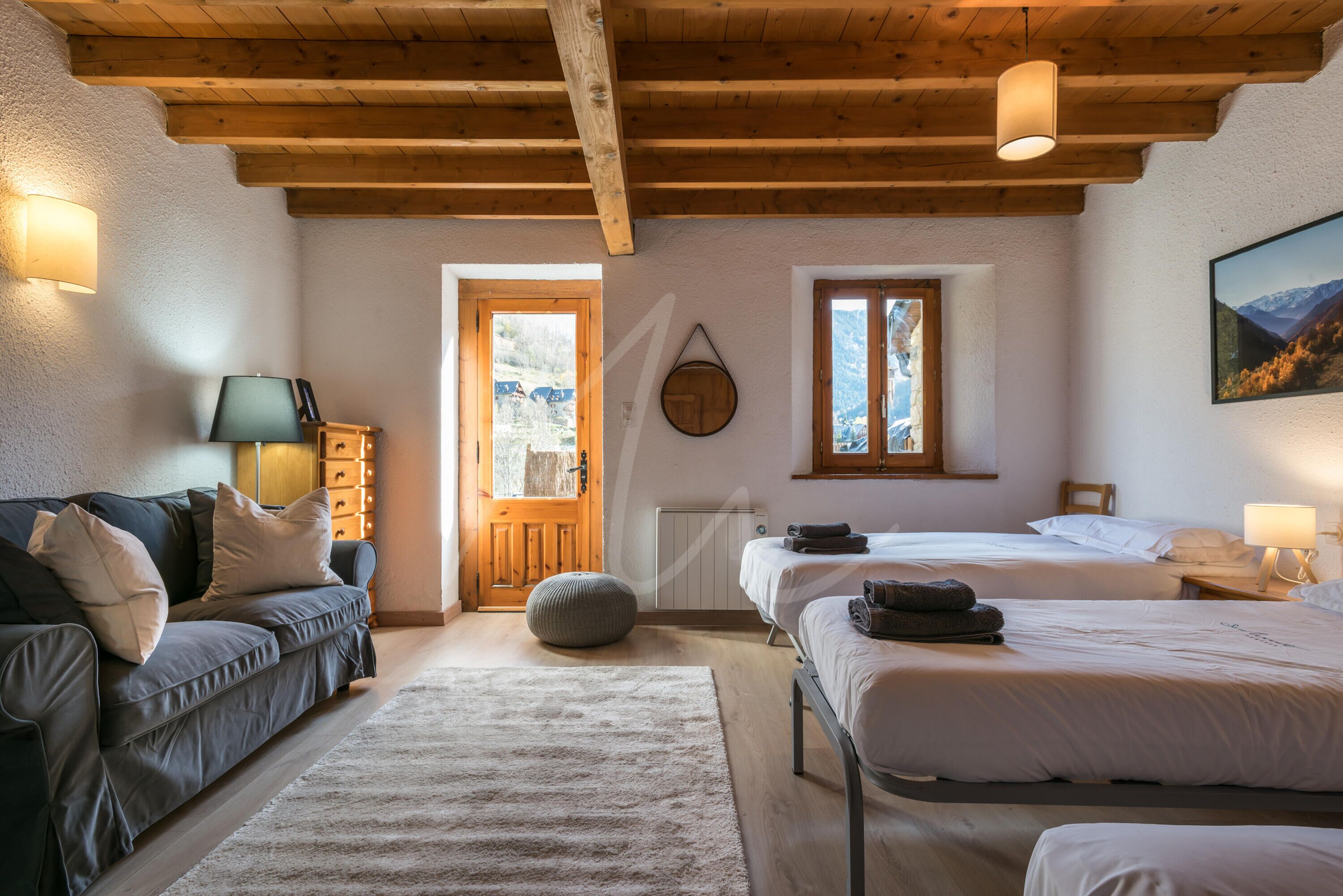 Una habitación acogedora con techo de madera, un sofá, dos camas, un radiador y vistas a las montañas a través de una ventana.