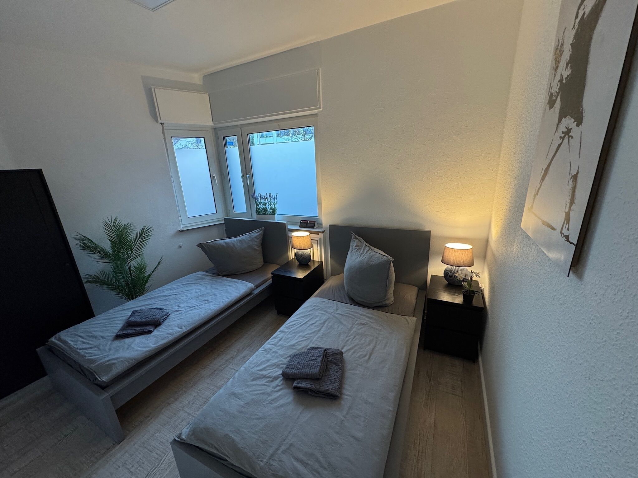 Ein kleines, modernes Schlafzimmer mit zwei Einzelbetten, einem Nachttisch mit Lampe, einem Fenster und einer Pflanze.