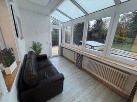 Um solário com sofá de couro preto, radiador e vista para o exterior.