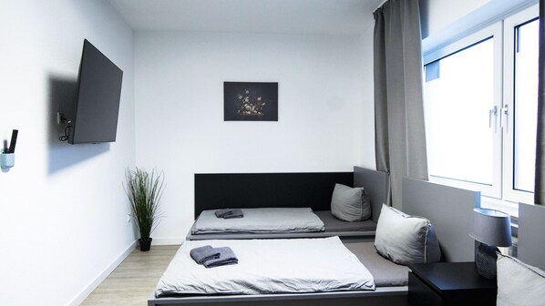 Een moderne slaapkamer met twee bedden, een flatscreen televisie, een potplant en een raam met gordijnen.