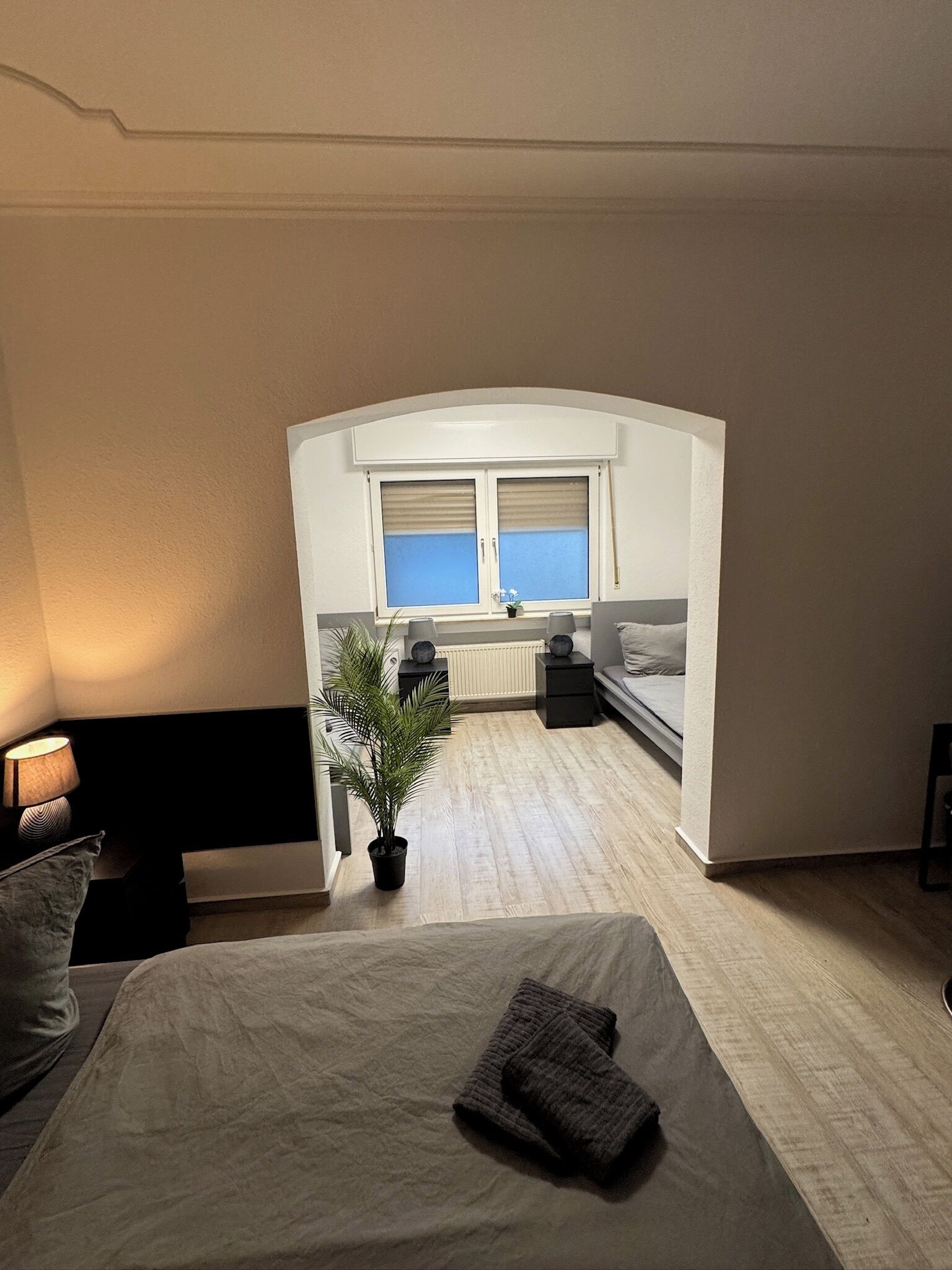 A bedroom with a bed, a nightstand, a lamp, a potted plant, a window, and a view of the kitchen.
