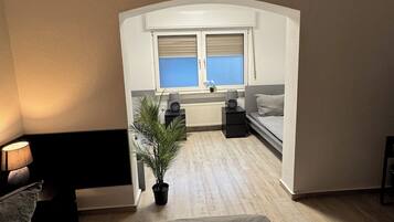 A bedroom with a bed, a nightstand, a lamp, a potted plant, a window, and a view of the kitchen.