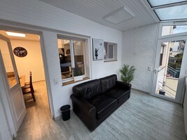 Quarto com sofá de couro preto, piso de madeira, parede branca com textura, janela com vista e uma pequena lixeira.