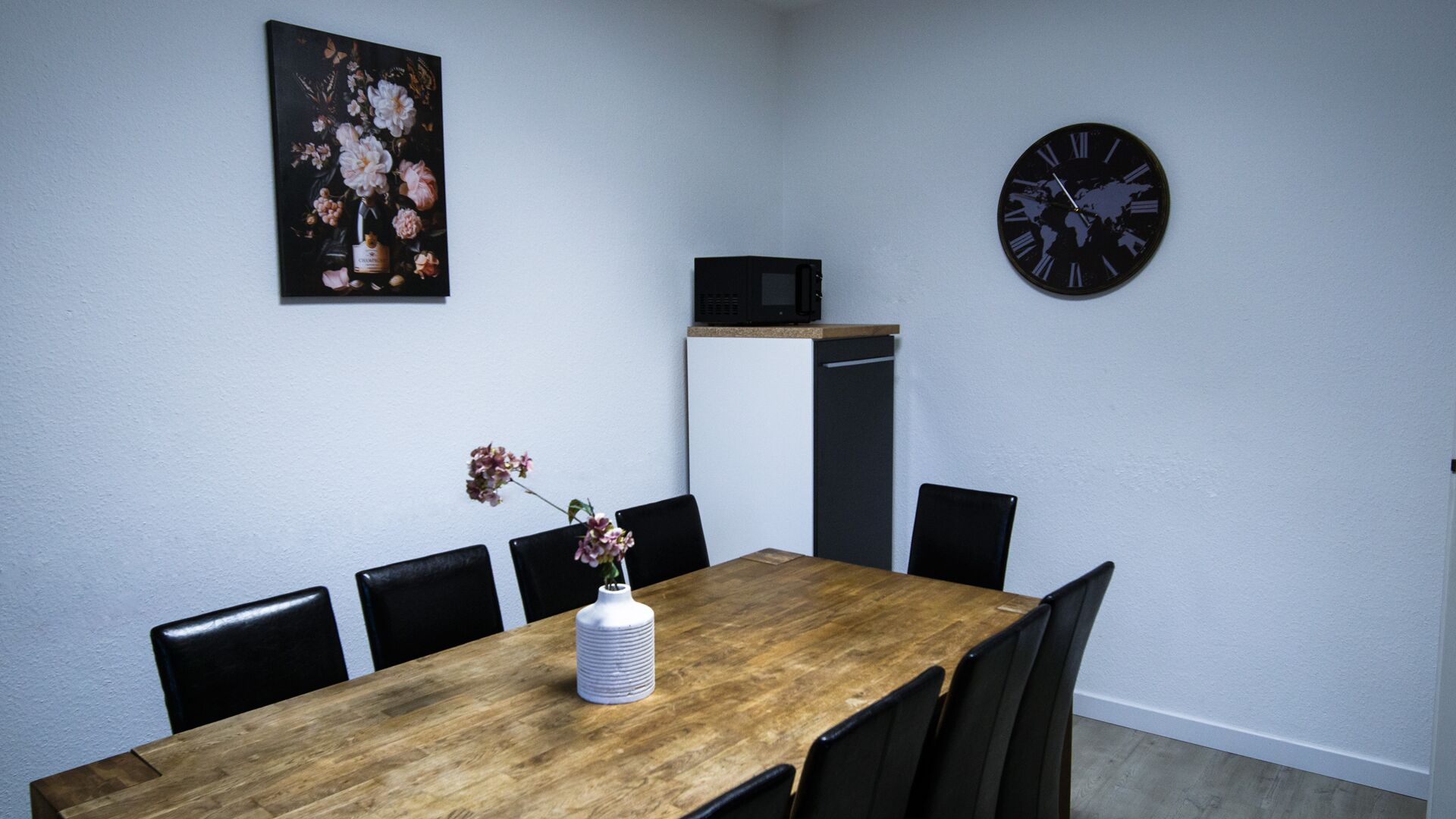 En matplats med ett träbord, svarta stolar, en klocka, en mikrovågsugn och en blomstermålning på väggen.