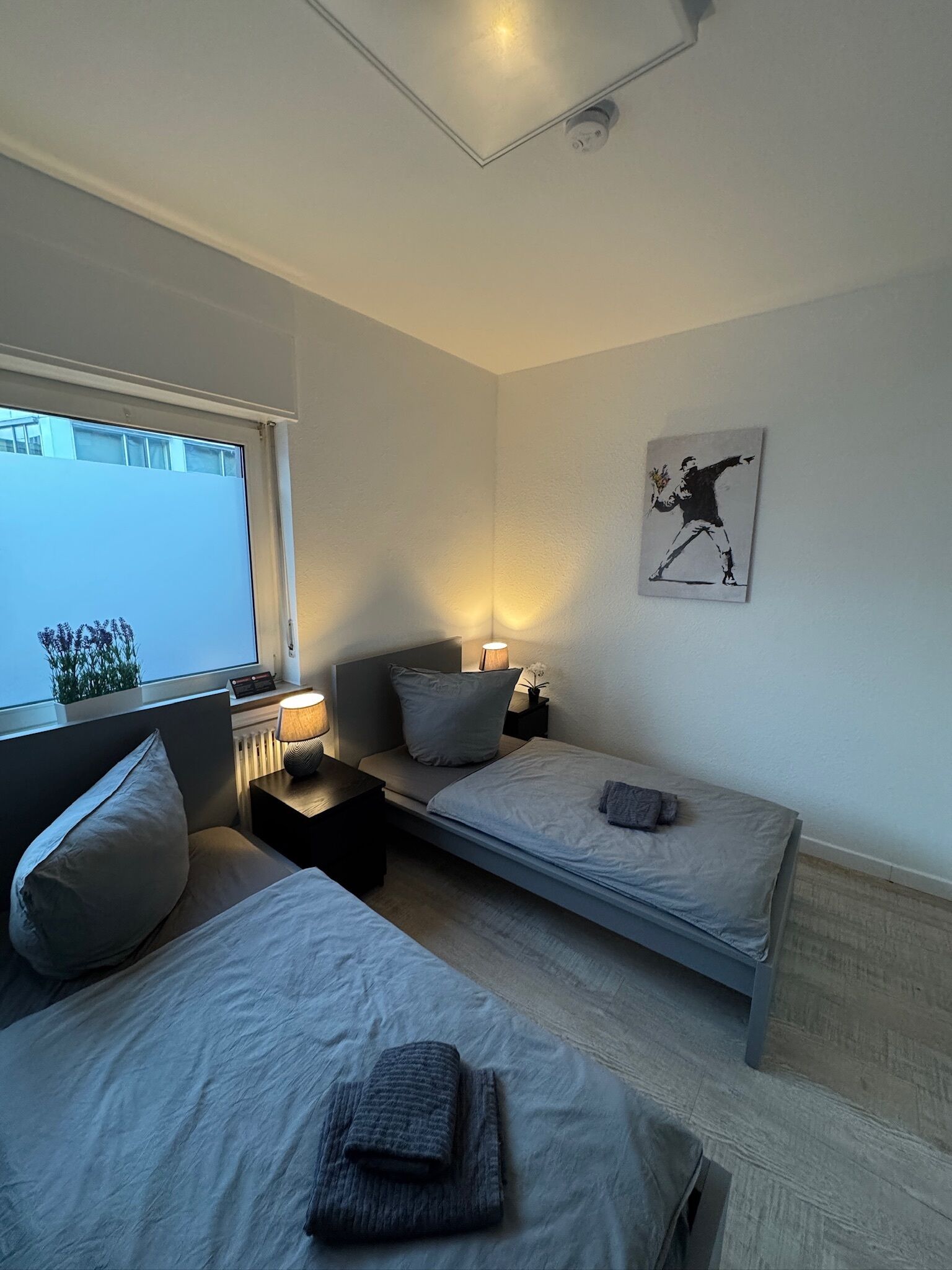 A bedroom with two single beds, bedside tables with lamps, a window, and a wall-mounted artwork.