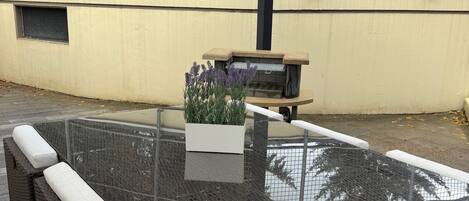 Eine Terrasse im Freien mit einem Glastisch, Korbstühlen mit Kissen, einem kleinen Pflanztopf mit Lavendel und einem Metallzaun im Hintergrund.