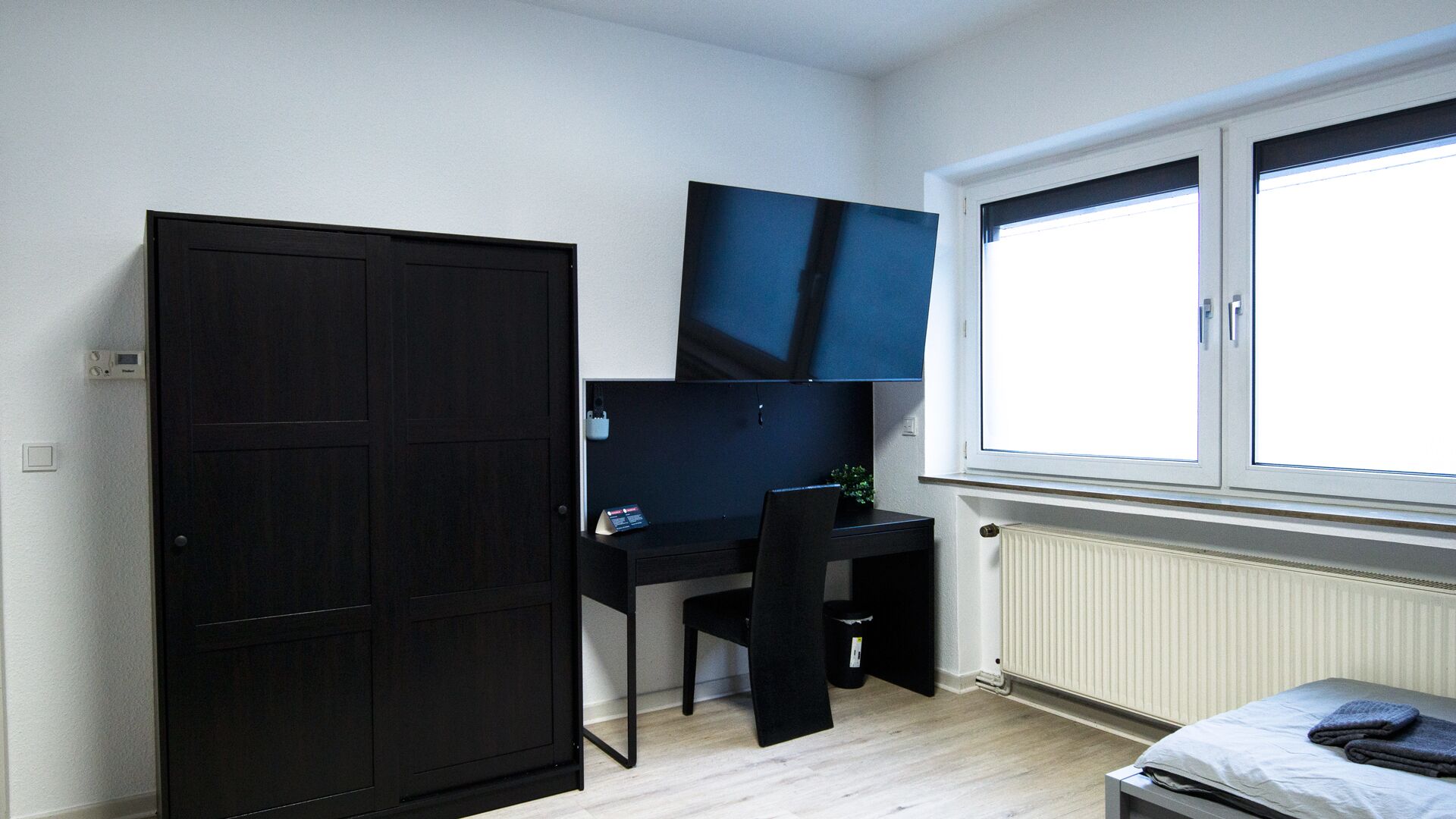 A room with a dark wardrobe, a desk with a chair, a bed, and a window with blinds.
