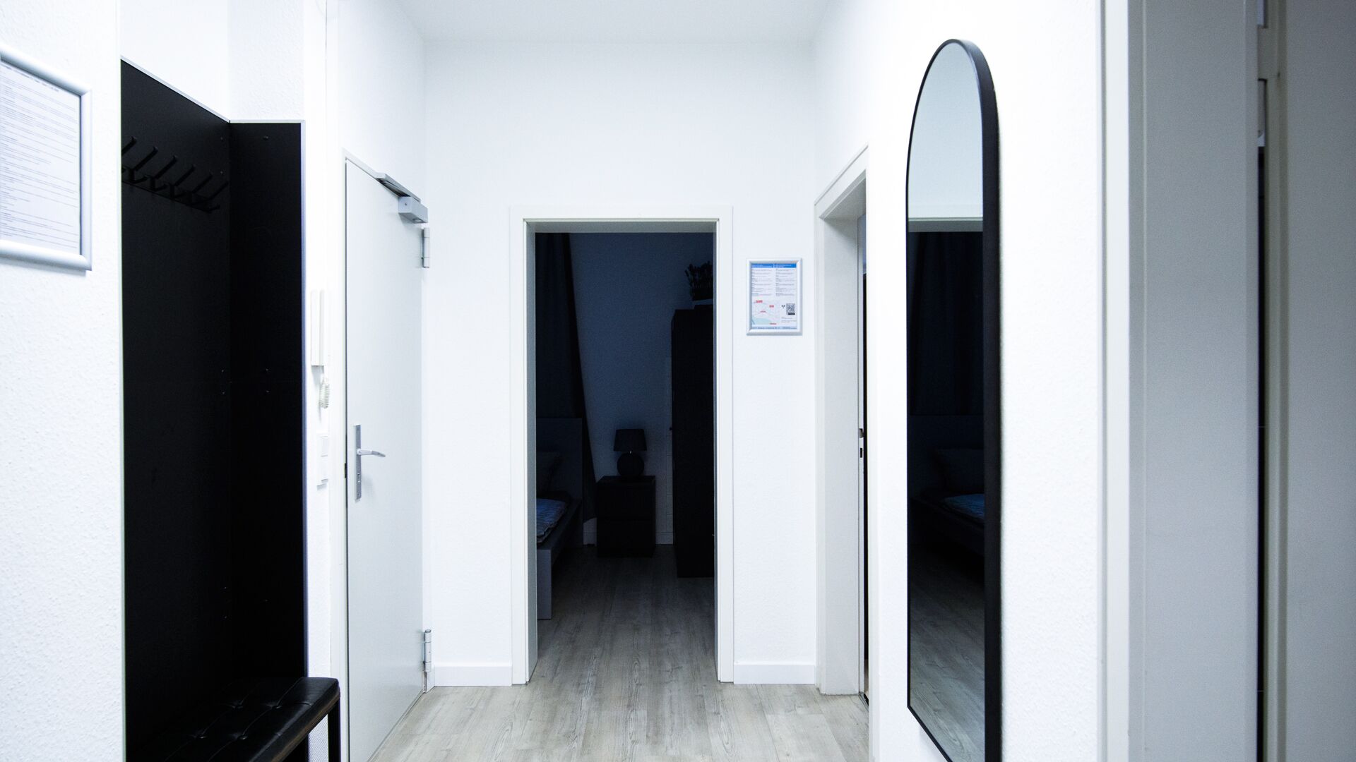 A hallway with a bed, a mirror, and a door leading to a dark room.