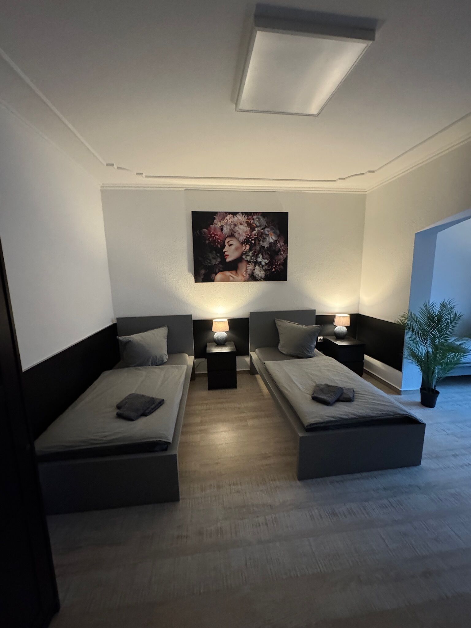 A modern bedroom with two beds, a nightstand with a lamp, a painting on the wall, and a potted plant.