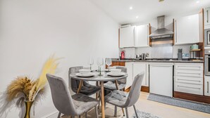 A modern kitchen with white cabinets, a stainless steel range hood, and a dining area with a round table and grey upholstered chairs. - The Cosy Spot- Contractor Friendly- Free Parking (Clapham)