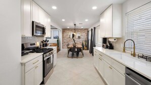 A modern kitchen with white cabinets, stainless steel appliances, and a dining area with a brick wall. - 6BD/3.5BA Modern Rustic Retreat | Yard, Pool & BBQ (Richardson)