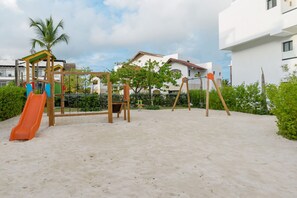 A playground with a slide and swings, surrounded by sand and greenery, with buildings in the background. - ONA | Terrace Picuzzi & Beach (Bavaro)
