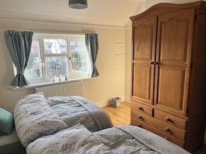A bedroom with a large wooden wardrobe, a bed with floral bedding, a window with curtains, and a radiator.