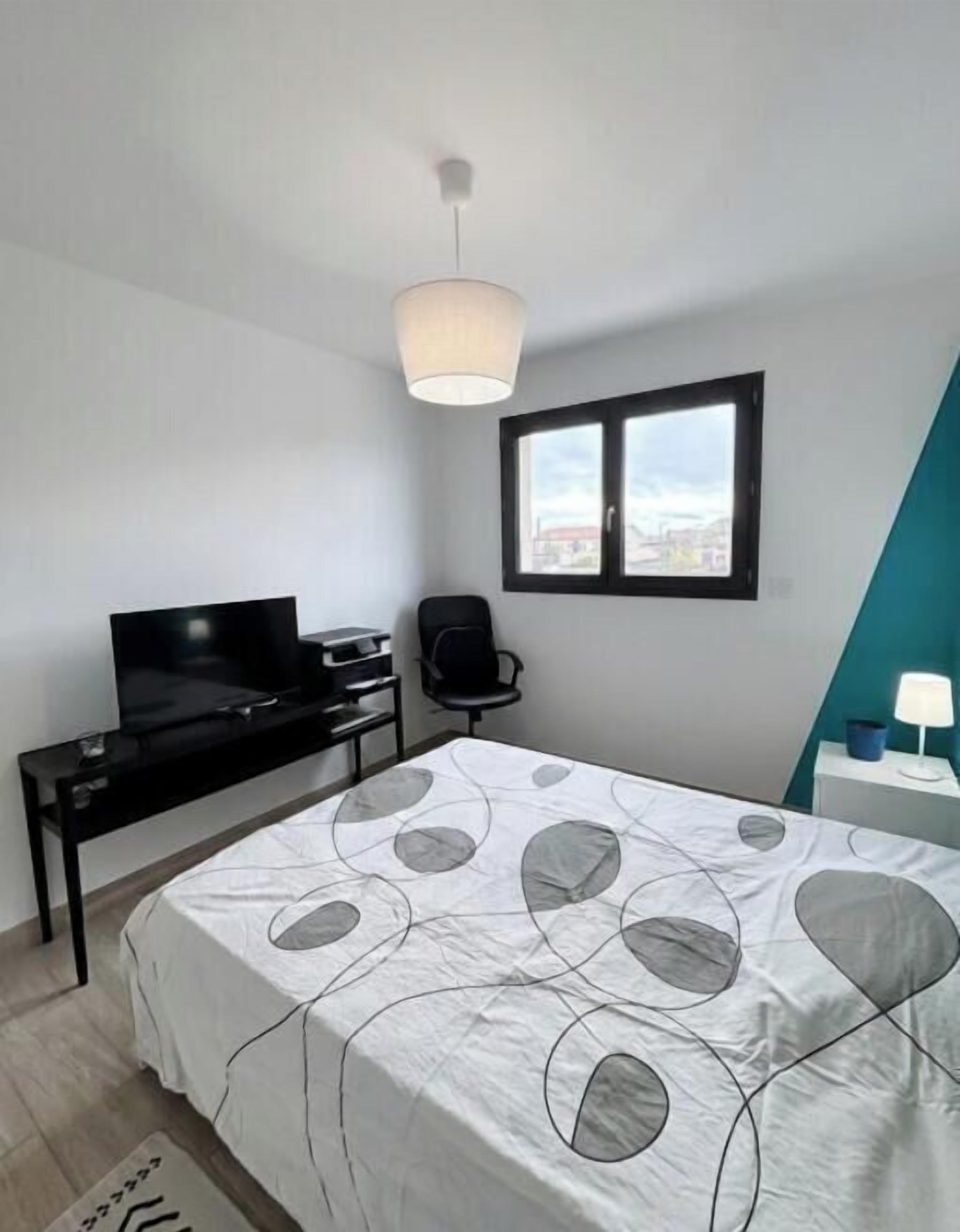 Un dormitorio con cama, escritorio con televisor, silla, ventana y lámpara.