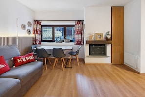 Una sala de estar moderna con un sofá gris, cojines rojos, una mesa de comedor con sillas, una chimenea y un ventanal con cortinas.