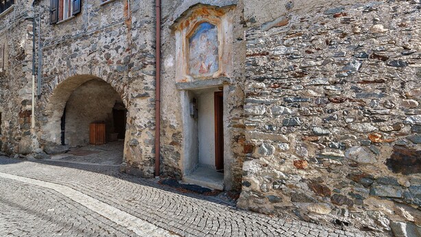 Ein Steinhaus mit Bogenportal, einem kleinen Fenster mit Fresko und einer gepflasterten Straße.