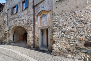 Un edificio de piedra con un arco de entrada, un pequeño ventanal con un fresco y una calle adoquinada.