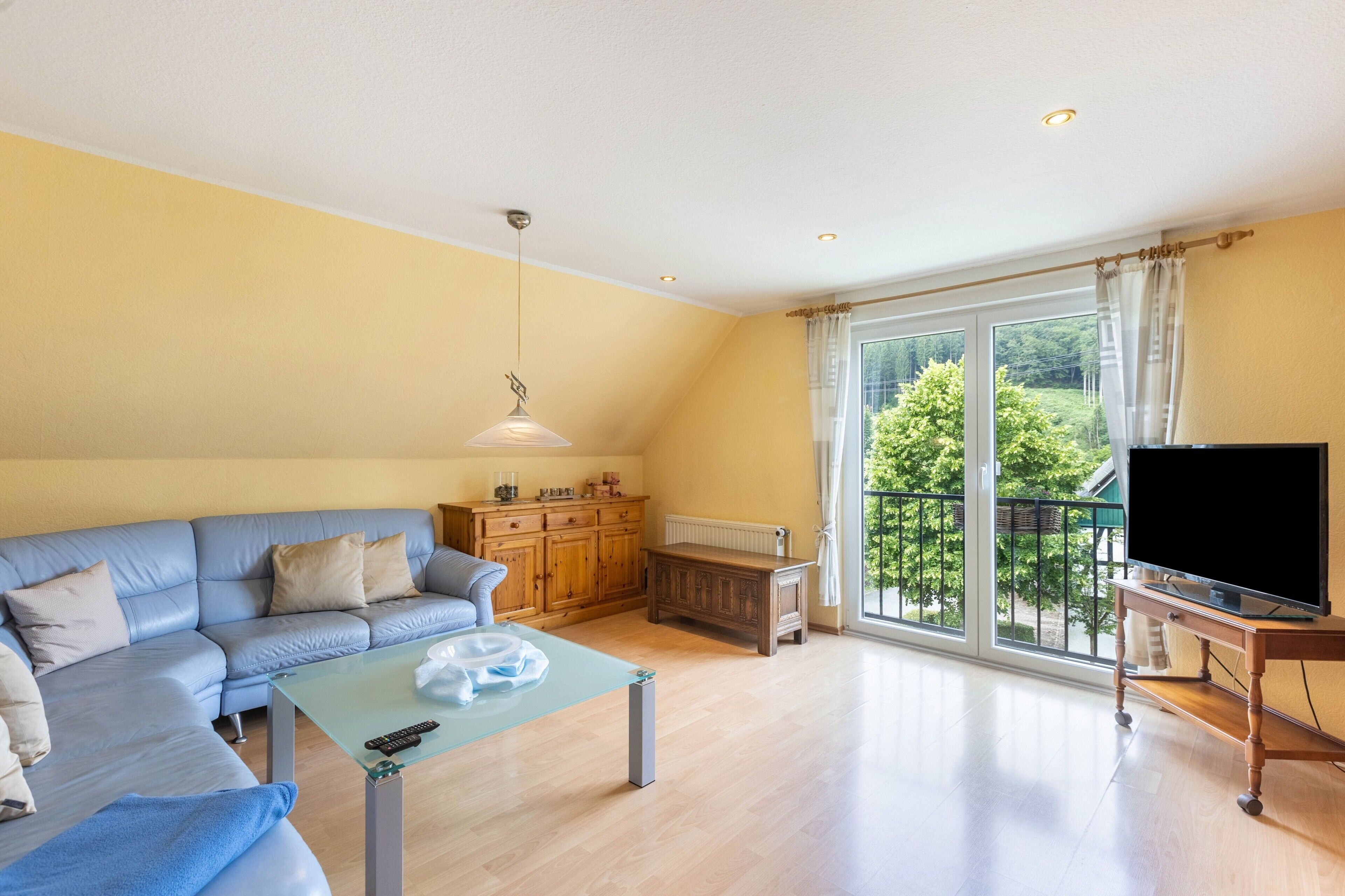 Ein Wohnzimmer mit einer Couch, einem Couchtisch, einem Fernseher auf einem Schrank, einer Holzkommode und einem großen Fenster mit Blick auf Grünflächen.