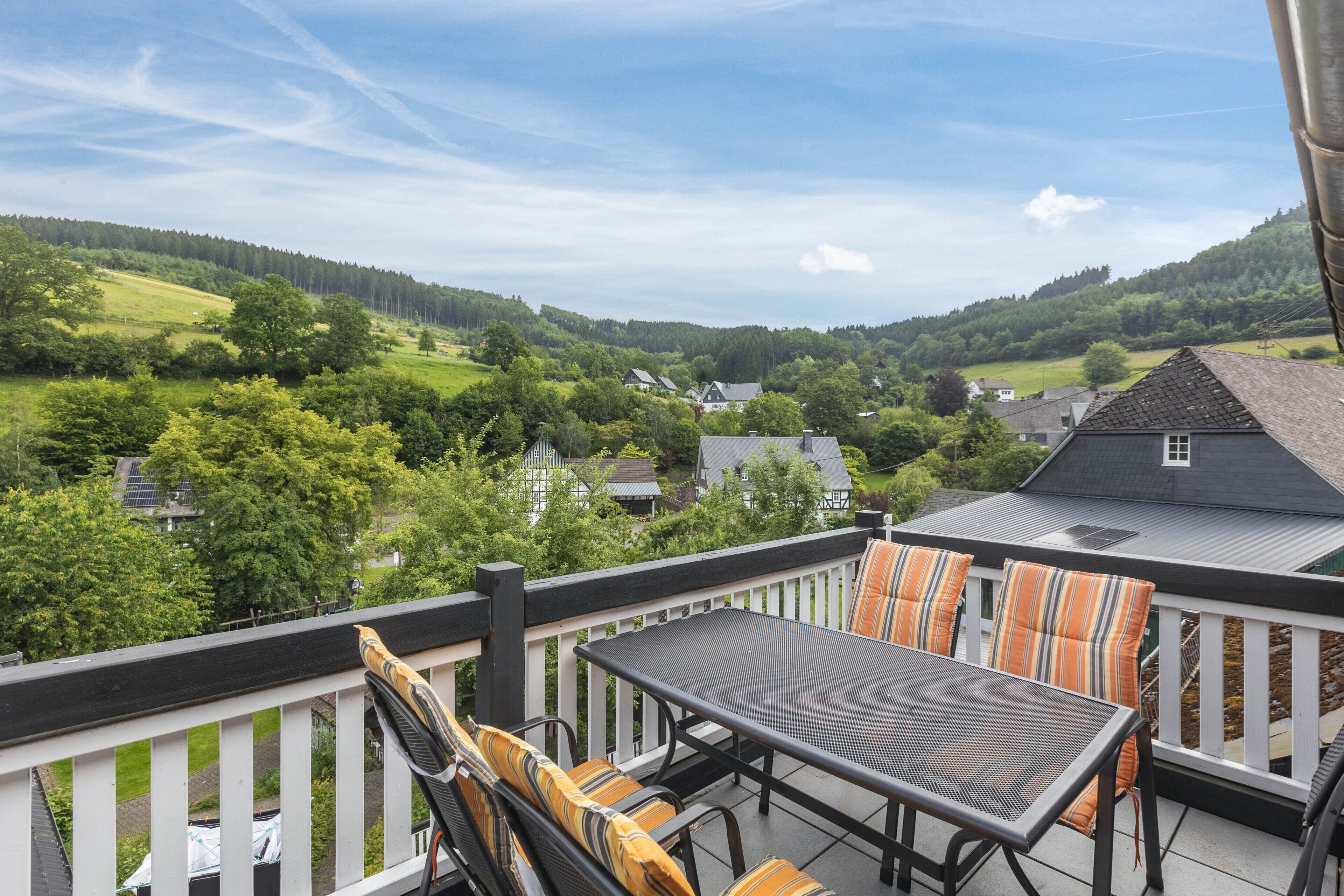 A balcony with a table and chairs, overlooking a green landscape with houses.