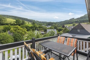 Un balcón con mesa y sillas, con vista a un paisaje verde con casas.