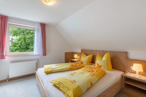 Ein Schlafzimmer mit einem hölzernen Kopfteil, einem Fenster mit roten Vorhängen, einem Heizkörper und einem Bett mit gelber Blumenbettwäsche.