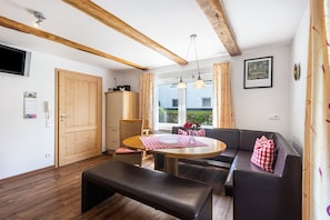 Ein Essbereich mit einem runden Tisch, Bänken und einer Couch. Eine Holztür, ein Fernseher und ein Fenster mit Vorhängen sind ebenfalls vorhanden.