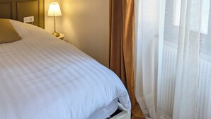 Cama bem arrumada com edredom branco e um travesseiro dourado. Abajur com cúpula branca sobre mesa de cabeceira de madeira. Janela com cortinas finas e brancas visível, permitindo a entrada de luz natural.