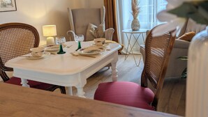 Uma área de refeições com uma mesa branca posta para dois, rodeada por cadeiras de vime. Uma pequena mesa redonda com um vaso de plantas secas é visível ao fundo.