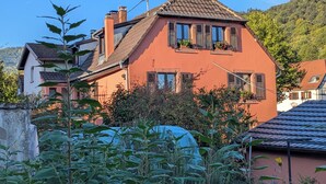 Uma casa de dois andares com telhado inclinado e persianas, cercada por vegetação e outras casas.