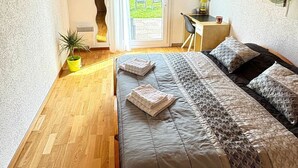 Een slaapkamer met een houten plafond, een bed met beddengoed met een patroon, een bureau met een stoel, een potplant en uitzicht door een raam.