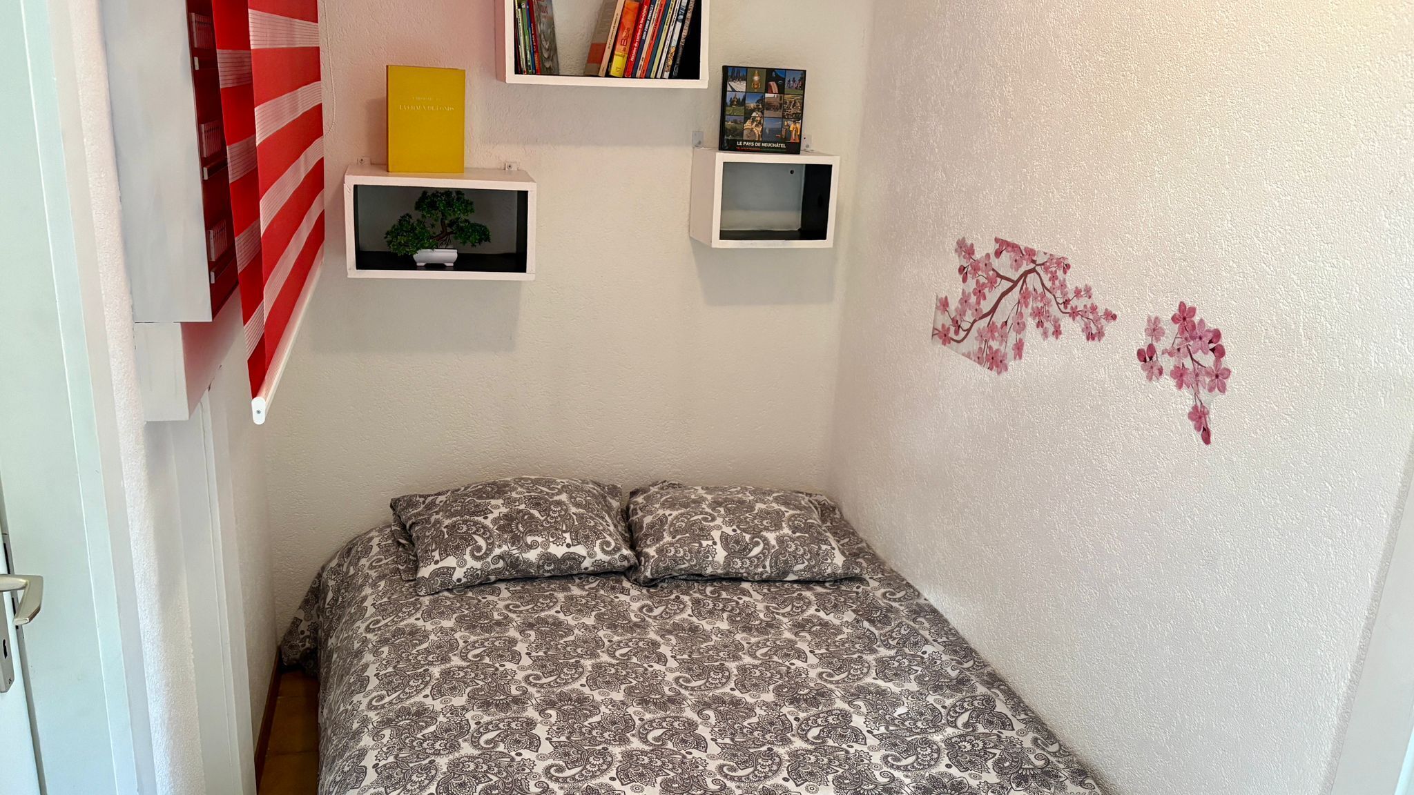 A bedroom with a patterned bedspread, two pillows, a small shelf with books, a framed picture, and a wall decal of flowers.