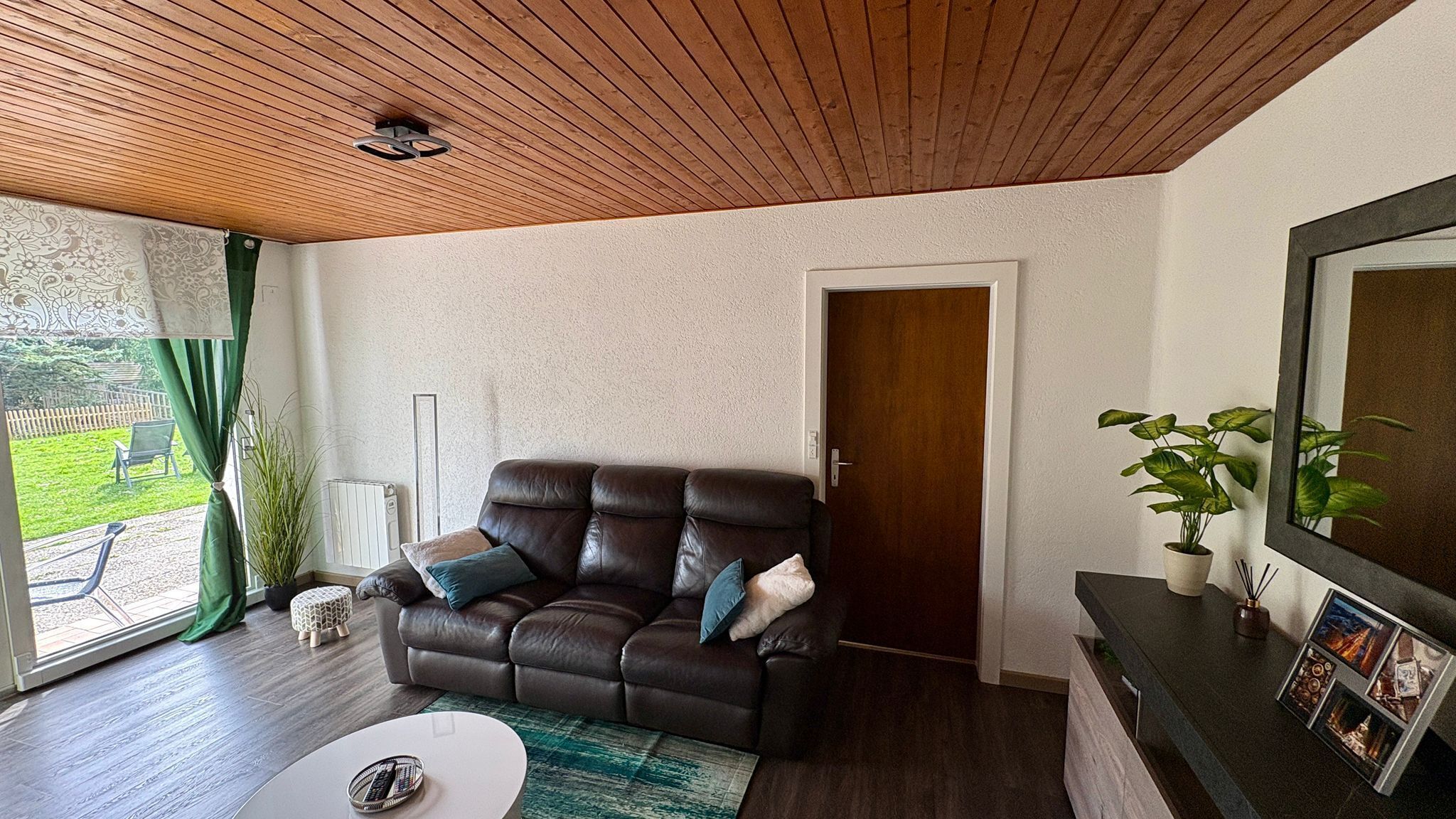 Een woonkamer met een leren bank, een klein wit tafeltje, een radiator, een deur en een groot raam met gordijnen.