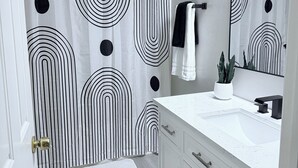 Ein Badezimmer mit weißem Waschtisch, einem schwarzen Wasserhahn, einer weißen Spüle, einem Spiegel und einem Duschvorhang mit einem schwarz-weißen abstrakten Muster.