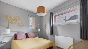 Ein Schlafzimmer mit Bett, Nachttischen, einer Hängeleuchte, einem Sessel und einem Fenster mit Vorhängen.