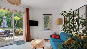 Ein modernes Wohnzimmer mit einem blauen Sofa, einem runden Holzschemel, einem Fernseher und einem großen Fenster mit Blick auf Grünflächen.