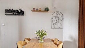 Ein Essbereich mit Holztisch, sechs Stühlen, einer Vase mit Blumen, einem Stadtbild-Kunstwerk, einem Regal mit Dekoration und einem gerahmten abstrakten Kunstwerk.