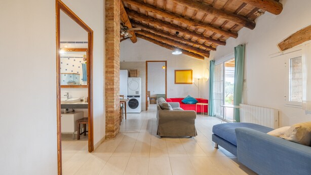 Uma sala de estar ampla com teto de madeira alto, um sofá azul, um sofá vermelho e uma cozinha ao fundo.