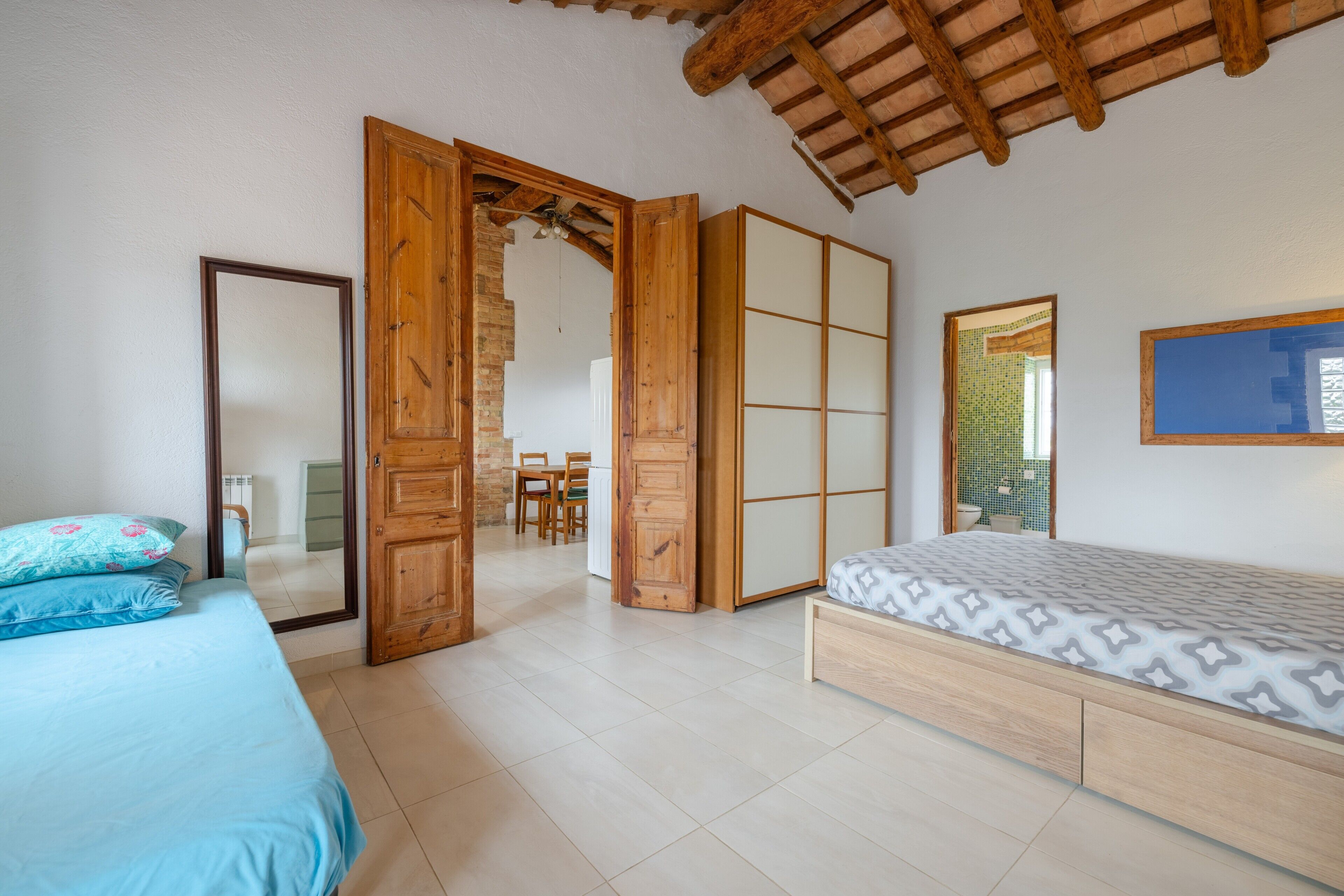 Une chambre à coucher comprenant un lit, une armoire en bois, une porte coulissante donnant accès à une salle de bain et un plafond avec des poutres apparentes.