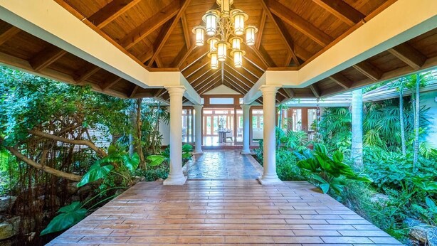 A covered walkway with wooden flooring and white pillars, surrounded by lush greenery and a view of a building in the distance.