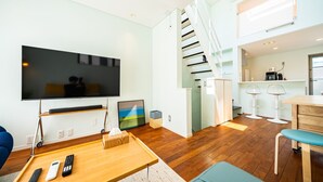 Un salon moderne avec un téléviseur à écran plat, une table basse en bois et un canapé. Un escalier mène à un niveau supérieur, et il y a un coin cuisine avec des tabourets de bar.