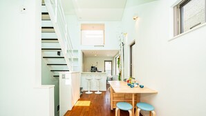 Un couloir étroit avec un escalier sur la gauche, un coin cuisine avec une table et des tabourets à manger sur la droite, et une fenêtre avec des stores.
