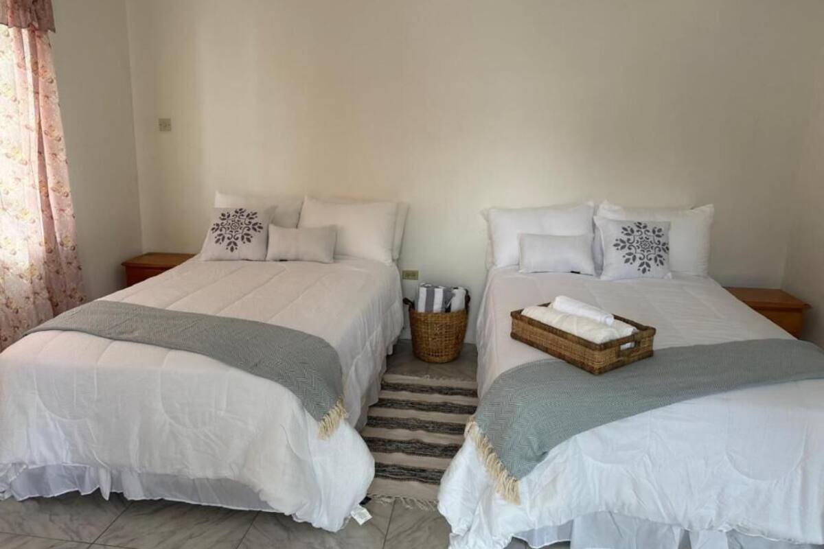 Una habitación con dos camas, cada una con ropa blanca y almohadas decorativas. Hay una cesta de mimbre en el suelo entre las camas.