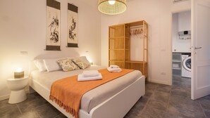 Un dormitorio con cama, mesitas de noche, una lámpara colgante, un armario de madera y una lavadora en la habitación contigua.